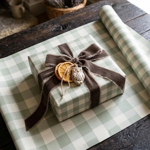 Op de afbeelding: Een vierkant cadeaupakket verpakt in saliegroen en wit vichy-ruitpapier, vastgebonden met een donkerbruin gerafeld fluwelen lint. Het pakket is versierd met twee gedroogde sinaasappelschijfjes en een klein wit keramieken uilornament.