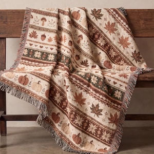 Könnte beinhalten: Eine gewebte Wohndecke mit einem sich wiederholenden Muster aus Kürbissen, Ahornblättern, Eicheln, Pilzen und kleinen Herzen in Creme-, Braun- und Dunkelgrüntönen. Die Decke hat eine strukturierte Oberfläche und ausgefranste Ränder.