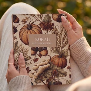 May include: A rectangular journal with a cream-colored cover featuring a botanical illustration of pumpkins, acorns, wheat stalks, and leaves in shades of brown and tan. A central rectangular label displays the text NORAH AUTUMN JOURNAL.