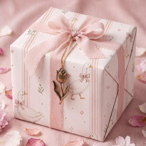 May include: A square gift box wrapped in white paper featuring a pattern of white geese wearing pink bows and vertical pink stripes. The box is tied with a wide pink ribbon and adorned with a gold-toned metal tulip charm on a delicate chain.