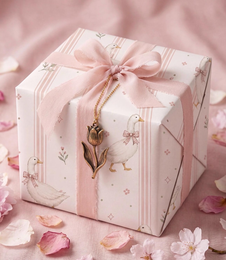 May include: A square gift box wrapped in white paper featuring a pattern of white geese wearing pink bows and vertical pink stripes. The box is tied with a wide pink ribbon and adorned with a gold-toned metal tulip charm on a delicate chain.