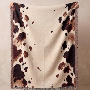 May include: A rectangular woven throw blanket featuring a cream-colored base with irregular dark brown and tan cowhide-style spots. The blanket has a textured appearance and is finished with decorative fringe along the two vertical side edges.