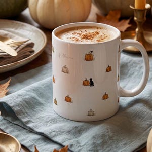 May include: A white ceramic mug filled with a latte topped with cinnamon, featuring a repeating pattern of small orange pumpkins, beige pumpkins, and a black cat. The name Katherine is printed in cursive script on the side of the mug.