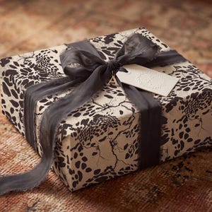Pode incluir: Uma caixa de presente quadrada embrulhada em papel creme com um padrão de leopardo abstrato preto. A caixa está atada com uma fita de seda cinzento-escura desfiada, com um pendente dourado em forma de letra A e uma etiqueta branca com coroa.
