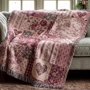 May include: A woven throw blanket featuring a patchwork design in shades of burgundy, cream, and muted pink. The pattern includes floral motifs, heart shapes, plaid squares, and toile-style scenes, finished with a fringed edge on all sides.