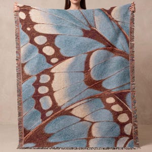 May include: A rectangular woven throw blanket featuring a large-scale graphic print of a butterfly wing. The design displays shades of light blue, cream, and dark brown with intricate vein patterns and a decorative fringed edge on all sides.