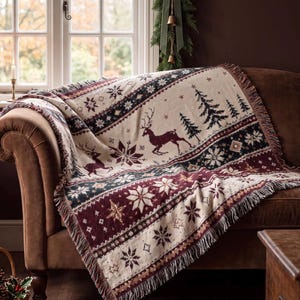 Puede incluir: Una manta tejida con una base de color crema y bandas horizontales en verde oscuro y burdeos. El patrón incluye motivos repetidos de renos, pinos y varios diseños de copos de nieve, rematado con un borde de flecos.