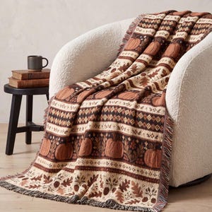 Op de afbeelding: Een geweven plaid met een herhalend patroon van oranje pompoenen, bruine eikenbladeren en geometrische ruitvormen in tinten crème, bruin en oranje. De deken heeft een rand met franjes en is gedrapeerd over een witte, getextureerde fauteuil.