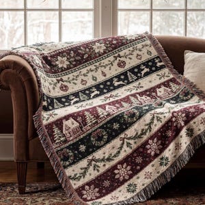Puede incluir: Una manta tejida con un patrón navideño de rayas horizontales en rojo oscuro, verde oscuro y crema. El diseño incluye ilustraciones de cabañas de invierno, pinos, renos, copos de nieve, flores de pascua y adornos navideños.