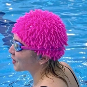 May include: A person wearing a bright pink, textured swim cap and blue-tinted swim goggles in a swimming pool. The person is wearing a black swimsuit. The water is a bright blue color.