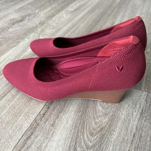 Könnte beinhalten: Ein Paar weinrote Keilabsatzschuhe. Die Schuhe haben ein Strickobermaterial mit einem kleinen herzförmigen Logo an der Seite. Der Keilabsatz ist etwa 5 cm hoch und besteht aus hellbraunem Material.