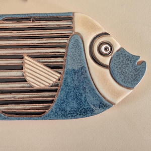 Könnte beinhalten: Keramische Fisch-Wanddekoration in Blau, Beige und Schwarz. Der Fisch hat einen strukturierten, gesprenkelten blauen Körper, einen beigen Kopf und ein Streifenmuster. Der Fisch ist etwa 30 cm lang.