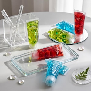 May include: Several clear squeeze tubes labeled Chiboky filled with colorful gel containing decorative elements like stars and red spheres. The tubes are arranged with a clear acrylic organizer, a glass tray, and a small mirror on a surface.