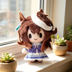May include: A soft toy doll with brown hair, a white headband, and a blue and white dress. The doll has a smiling face and is decorated with a blue gem. The doll is on a windowsill with potted plants.