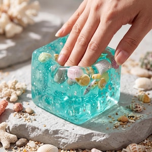 May include: A turquoise resin cube, filled with seashells and glitter, with a hand touching it. The cube is square with rounded edges. The image shows the cube surrounded by sand, coral, and more seashells.