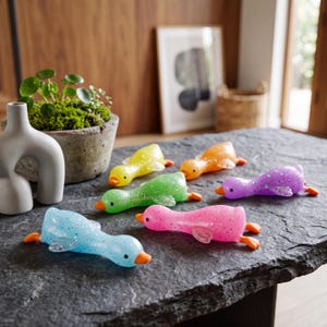 Puede incluir: Seis juguetes de pato brillantes y coloridos en tonos de azul, rosa, morado, verde, amarillo y naranja. Cada pato tiene pico y patas naranjas. Los juguetes están colocados sobre una superficie de piedra gris oscura.