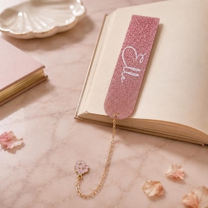 Puede incluir: Un marcapáginas rectangular de purpurina rosa con el nombre Elle en letras cursivas blancas. El marcapáginas está unido a una fina cadena de metal dorado que termina en un pequeño dije con forma de flor rosa claro y pétalos blancos.
