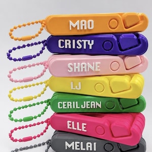 Puede incluir: Una pila de siete llaveros rectangulares de plástico en naranja, morado, rosa claro, amarillo, verde, rosa fuerte y gris. Cada llavero presenta un nombre blanco en relieve, un recorte circular y una cadena de bolas a juego.
