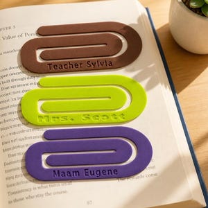 Puede incluir: Tres marcapáginas grandes y planos con forma de clip en colores marrón, verde lima y morado. Cada marcapáginas presenta texto en relieve: el marrón dice Teacher Sylvia, el verde Mrs. Scott y el morado Maam Eugene.