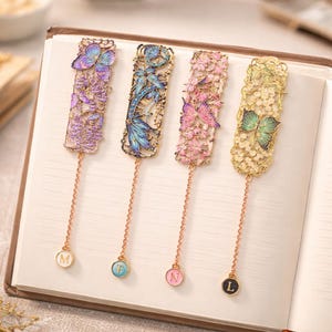 Puede incluir: Cuatro marcadores de metal decorativos con diseños de mariposas y flores. Cada marcador tiene una combinación de colores diferente: morado, azul, rosa y verde. Cada marcador tiene una cadena con un dije con una letra. Los marcadores están sobre las páginas de un libro.