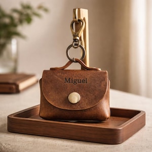 Può includere: Un astuccio in pelle marrone con il nome "Miguel" inciso sul davanti. L'astuccio è attaccato a un portachiavi e moschettone in metallo ed è esposto su un vassoio di legno. Ha una chiusura a bottone.