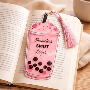 Pink boba bookmark with shameless smut lover text placed on open book cute romance reader accessory