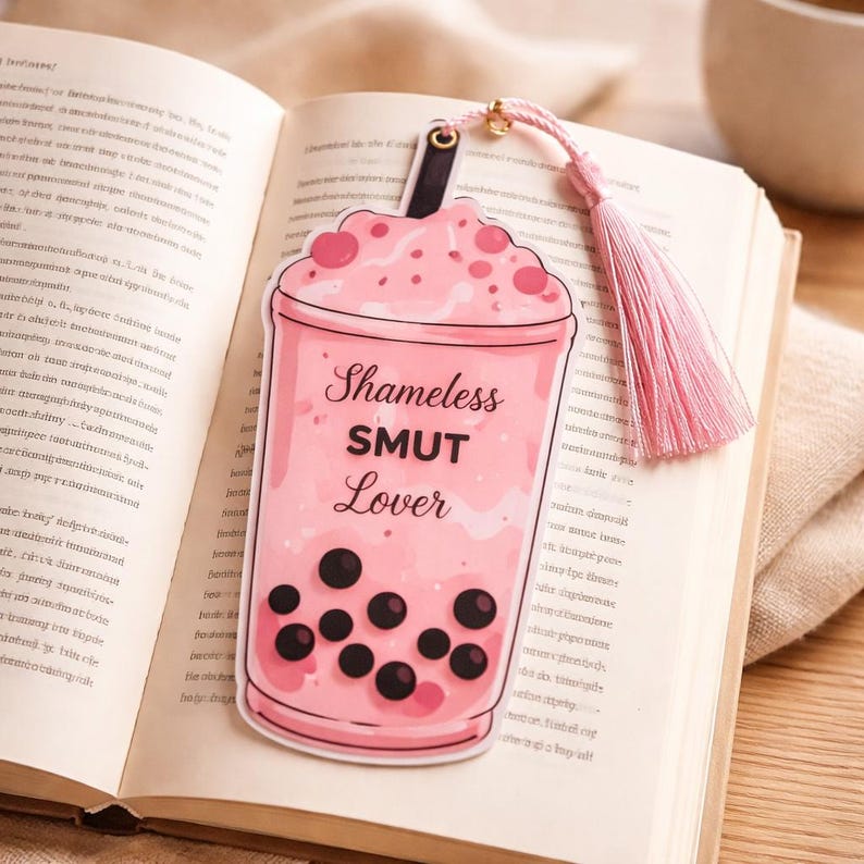 Pink boba bookmark with shameless smut lover text placed on open book cute romance reader accessory