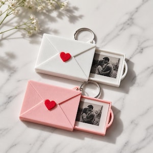 Pode incluir: Dois porta-chaves em forma de envelope, um branco e outro rosa claro, cada um com um pequeno coração vermelho na aba frontal. Um anel de metal está preso a cada um. Uma pequena fotografia quadrada a preto e branco de um casal é extraída.