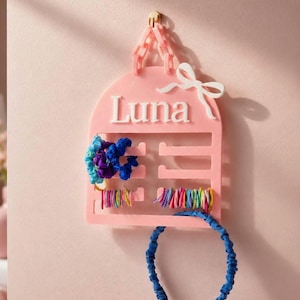 May include: A pink arched wall-mounted organizer featuring the name Luna in white lettering, a white bow accent, and a pink chain for hanging. The organizer holds several blue scrunchies, a row of colorful hair ties, and a blue fabric headband.