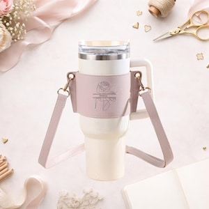 May include: A cream-colored tumbler with a handle and a clear lid, featuring a light pink leather sleeve. The sleeve is adorned with a floral design and the name "Jessica". A matching pink strap is attached for carrying.