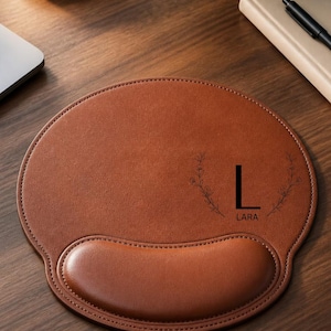 Pode incluir: Um mousepad de couro castanho com apoio para o pulso. A almofada tem uma borda costurada e apresenta a letra "L" com o nome "LARA" abaixo, juntamente com um design floral. A almofada está em uma mesa de madeira.