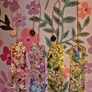 Four custom metal bookmarks with butterfly-floral designs and initial A charms on gold chains, a perfect gift for readers.