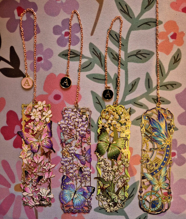 Four custom metal bookmarks with butterfly-floral designs and initial A charms on gold chains, a perfect gift for readers.
