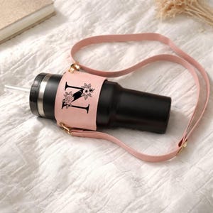 Puede incluir: Un vaso negro con pajita, con una funda de cuero rosa con la letra "N" y detalles florales. El vaso tiene una correa rosa a juego para llevar.