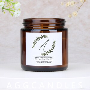 May include: A brown glass candle jar with a white label featuring a green leaf design and the letter "A". The label includes text that reads "Happy Birthday Richard!" and "Sandalwood rose" scent. The burn time is 16 hours.