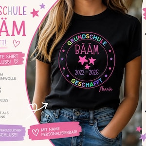 Op de afbeelding: Zwart T-shirt met een kleurrijke afbeelding met de tekst "GRUNDSCHULE BÄÄM GESCHAFFT!" met sterren en de jaren 2022-2026. Het shirt wordt getoond op een persoon die een spijkerbroek draagt. Extra kleuren zijn: zwart, wit en marine.