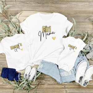 May include: White t-shirts and a onesie with the text "Girl Mama" and "Mama's Girl" in gold and black lettering. The image includes denim shorts, white sneakers, and baby shoes, arranged on a wooden surface with greenery.