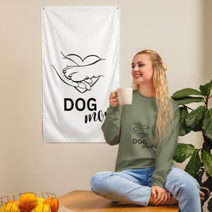 May include: A white flag with a black graphic of a dog paw and a heart, with the words "DOG mom" in black. A woman is wearing a green long-sleeve shirt with the same graphic and text. She is holding a white mug.