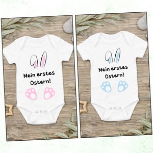 Puede incluir: Dos bodies blancos para bebé con diseños de Pascua. Uno presenta orejas y huellas rosas, el otro azules. Ambos tienen el texto "Mein erstes Ostern!" en negro, celebrando la primera Pascua de un bebé.