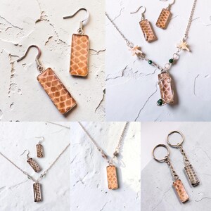 May include: A collection of jewellery featuring rectangular pendants with a tan, scale-like pattern. The set includes drop earrings with silver hooks, necklaces with delicate silver chains and small accents, and two keychains with silver rings.