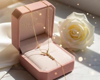 May include: A gold necklace with a Y-shaped pendant, adorned with small, sparkling stones, rests inside a pink velvet jewelry box. A white rose is in the background. The box is open, revealing the necklace against a soft, light pink interior.