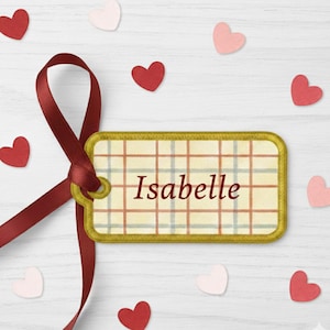 K&ouml;nnte beinhalten: Ein rechteckiges Geschenk-Etikett mit dem Namen "Isabelle" in Burgunderrot. Das Etikett hat einen goldenen Rand und ein kariertes Muster in Beige, Rot und Blau. Ein burgunderfarbenes Band ist an dem Etikett befestigt, und rote und rosa herzf&ouml;rmige Konfetti sind verstreut.
