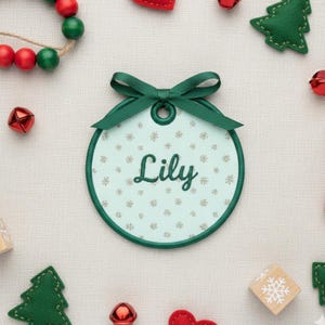 K&ouml;nnte beinhalten: Ein runder Weihnachtsschmuck mit dem Namen "Lily" in gr&uuml;ner Schrift gestickt. Der Schmuck hat einen hellblauen Hintergrund mit Schneeflockenmustern und einen gr&uuml;nen Rand. Oben ist ein gr&uuml;nes Band befestigt. Festliche Weihnachtsdekorationen umgeben den Schmuck.