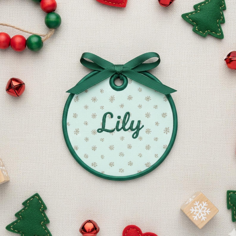 K&ouml;nnte beinhalten: Ein runder Weihnachtsschmuck mit dem Namen "Lily" in gr&uuml;ner Schrift gestickt. Der Schmuck hat einen hellblauen Hintergrund mit Schneeflockenmustern und einen gr&uuml;nen Rand. Oben ist ein gr&uuml;nes Band befestigt. Festliche Weihnachtsdekorationen umgeben den Schmuck.