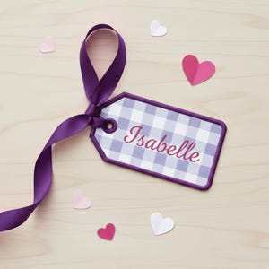 K&ouml;nnte beinhalten: Ein lila Vichy-Etikett mit dem Namen "Isabelle" in Schreibschrift, gebunden mit einem passenden lila Band. Papierherzausschnitte in Rosa und Wei&szlig; sind auf einer hellen Holzoberfl&auml;che verstreut.
