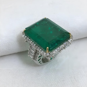 May include: A large emerald-cut green gemstone ring with a halo of small, clear gemstones. The ring has a silver band with additional small gemstones and gold accents on the corners of the emerald. The ring is set on a white surface.