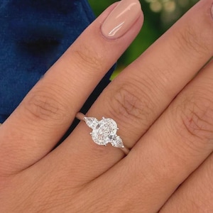 May include: A close-up of a diamond ring with a silver band. The ring features a large oval-cut diamond in the center, flanked by two smaller pear-shaped diamonds. The ring is set on a finger with a neutral nail polish.