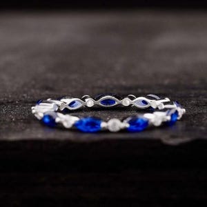 以下が含まれることがあります： 青と透明な宝石が交互に配置されたシルバーリング。青い宝石は楕円形で、透明な宝石は丸いです。リングは暗い背景に配置され、宝石の輝きを際立たせています。