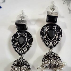 May include: A pair of silver-toned earrings with black gemstone accents. Each earring features a square top, a teardrop-shaped center with a black stone, and a bell-shaped bottom with dangling white beads. The design has an ornate, vintage aesthetic.