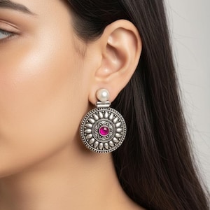 Puede incluir: Un pendiente plateado con un diseño circular con una piedra rosa central y un acento de perla. El pendiente tiene un patrón detallado, similar a una flor, alrededor de la piedra rosa. El pendiente se lleva en la oreja.